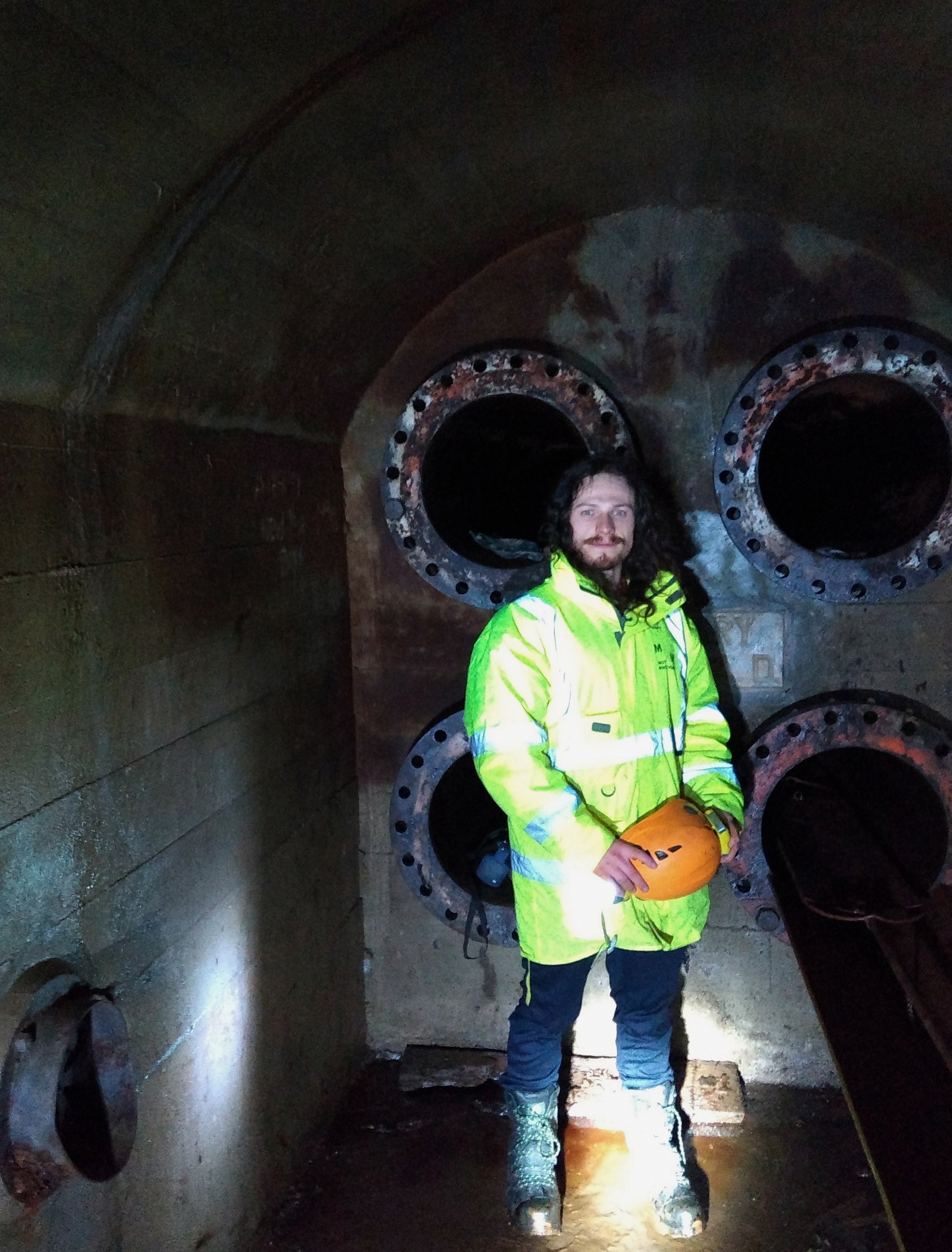 Tommy in a hi-vis jacket holding a pumpkin helmet, inside a tunnel lined with rusty pipe flanges